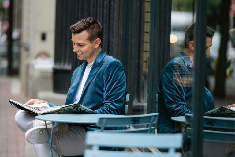 Matt Fraser in a blue jacket sits at an outdoor café table, reading a book and smiling, with his reflection in the nearby window.