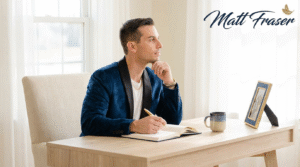 Matt Fraser, in a blue jacket, sits at a desk writing in a notebook, with a mug and framed picture beside him. His name appears onscreen.