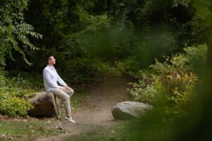 Matt Fraser in a white shirt and beige pants sits on a large rock along a dirt path surrounded by dense green foliage in a wooded area.