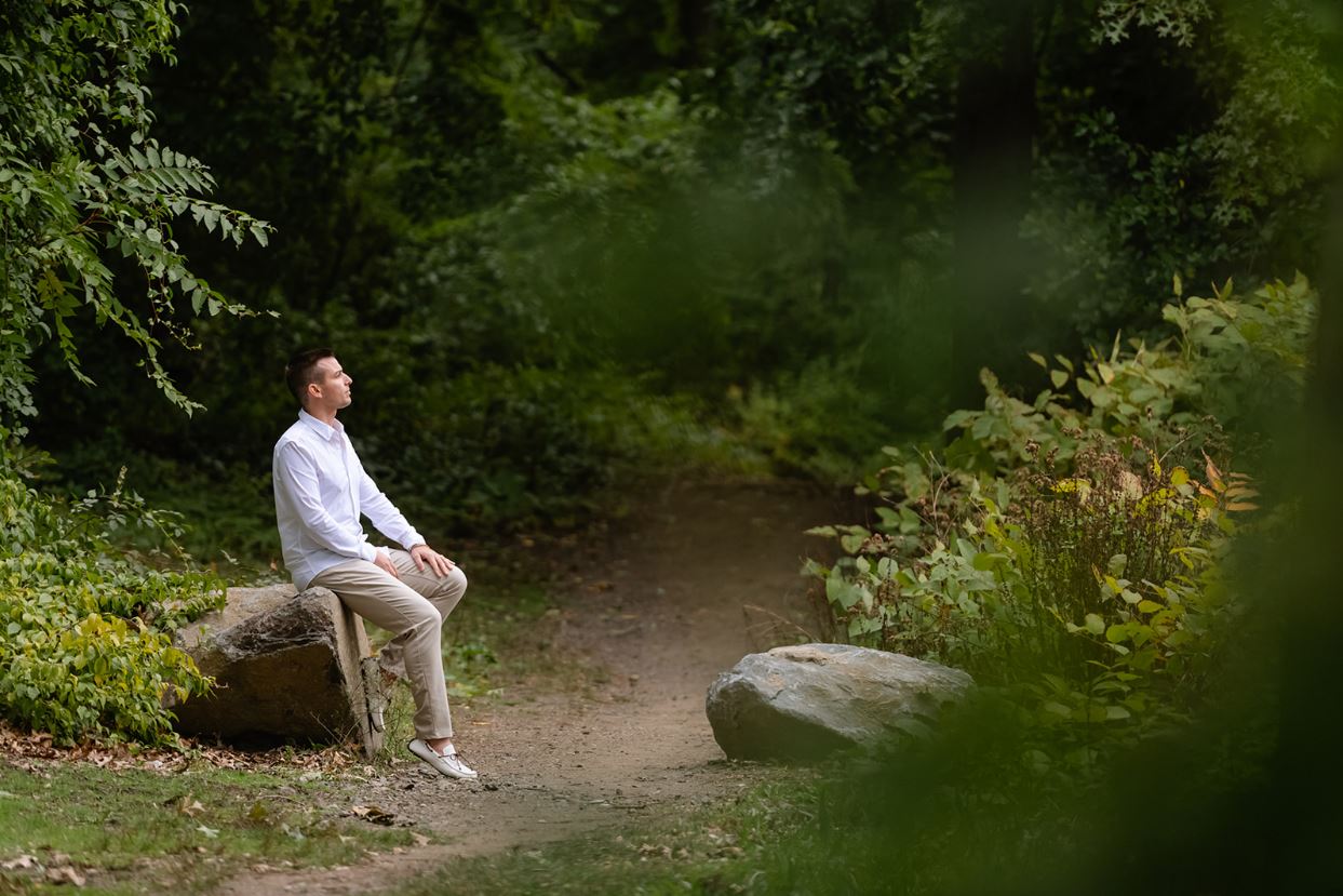 Matt Fraser in a white shirt and beige pants sits on a large rock along a dirt path surrounded by dense green foliage in a wooded area.