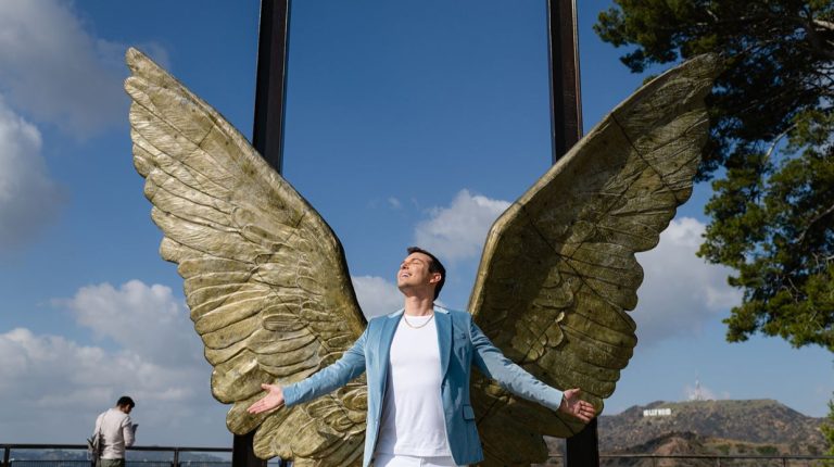 Matt Fraser stands in front of large metallic wing sculptures with arms outstretched, blue sky and the Hollywood sign in the background.