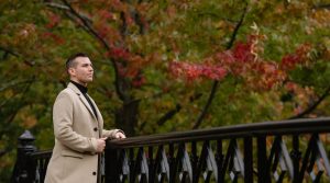 Matt Fraser in a beige coat stands at a black railing, looking thoughtfully into the distance with autumn trees behind him.
