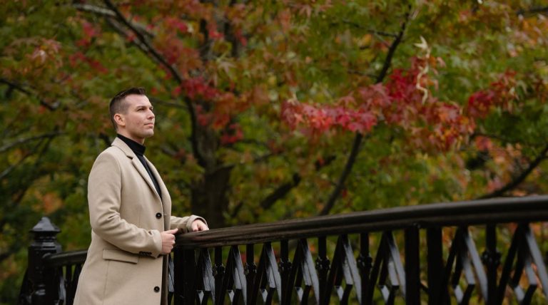 Matt Fraser in a beige coat stands at a black railing, looking thoughtfully into the distance with autumn trees behind him.