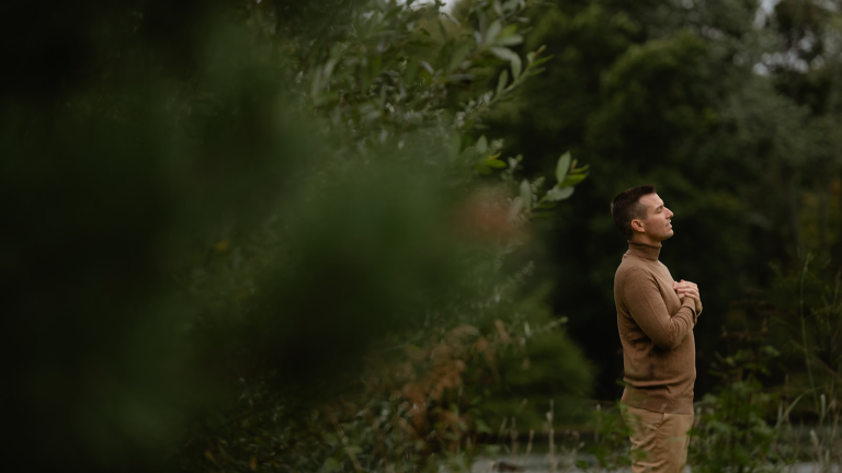 Matt Fraser in a brown turtleneck stands outdoors with hands clasped to his chest, surrounded by lush green foliage.