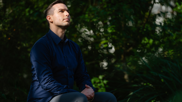 Matt Fraser in a dark blue shirt sits outdoors with hands clasped, looking upward, surrounded by greenery and dappled sunlight.