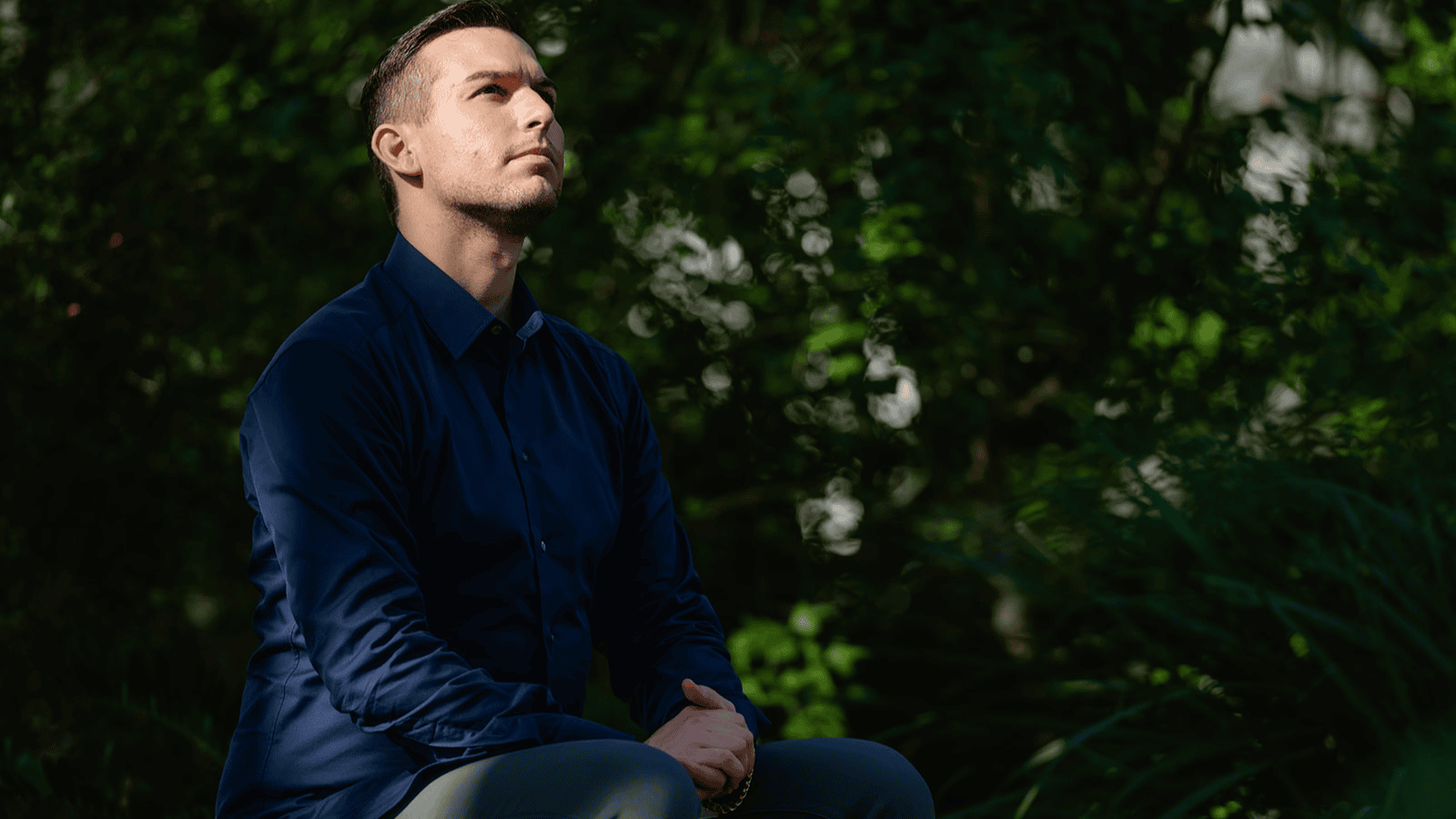 Matt Fraser in a dark blue shirt sits outdoors with hands clasped, looking upward, surrounded by greenery and dappled sunlight.