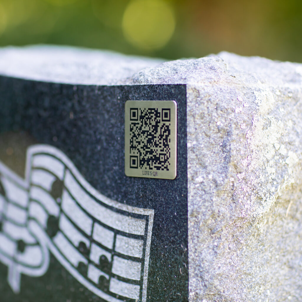 A metal QR code plaque labeled "LIFE'S QR" is fixed to a stone surface beside a carved musical staff design.