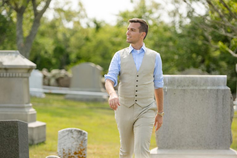 Matt Fraser in a light-colored vest and pants walks through a sunny cemetery, surrounded by headstones and greenery.