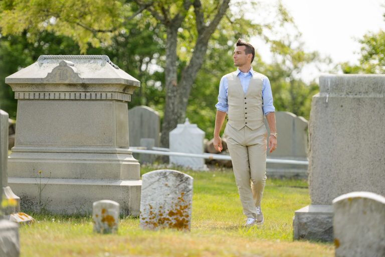 Psychic Medium Matt Fraser, dressed in a light vest and pants, walks through a cemetery with gravestones and greenery visible in the background.