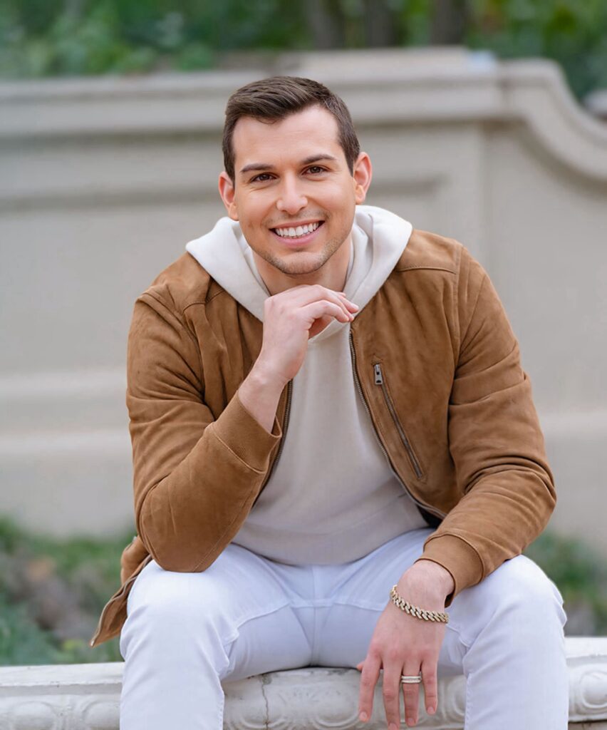 Matt Fraser, in a tan suede jacket and white pants, sits outdoors on a bench, smiling at the camera with his hand resting near his chin.
