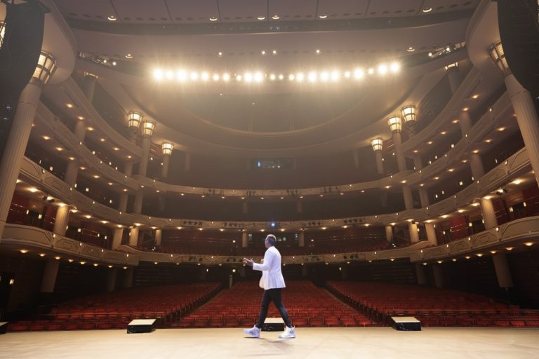 Matt Fraser in a white jacket stands alone on a well-lit theater stage, facing an empty auditorium with balconies and red seats.