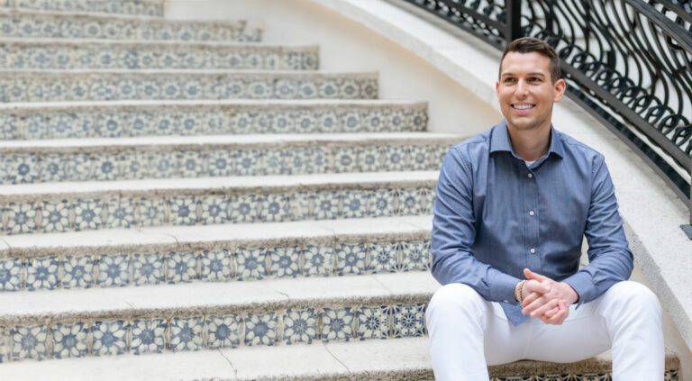 Matt Fraser, wearing a blue shirt and white pants, sits on patterned steps, smiling and looking to his left.