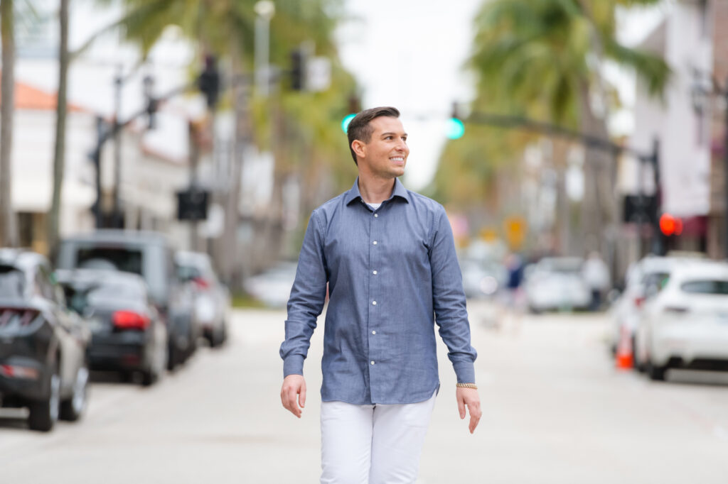 Matt Fraser, wearing a blue button-up shirt and white pants, walks down a street with trees and cars in the background.