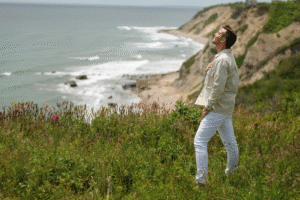 Matt Fraser, in a light shirt and white pants, stands on a grassy cliff by the ocean, looking upward with one hand on his chest.