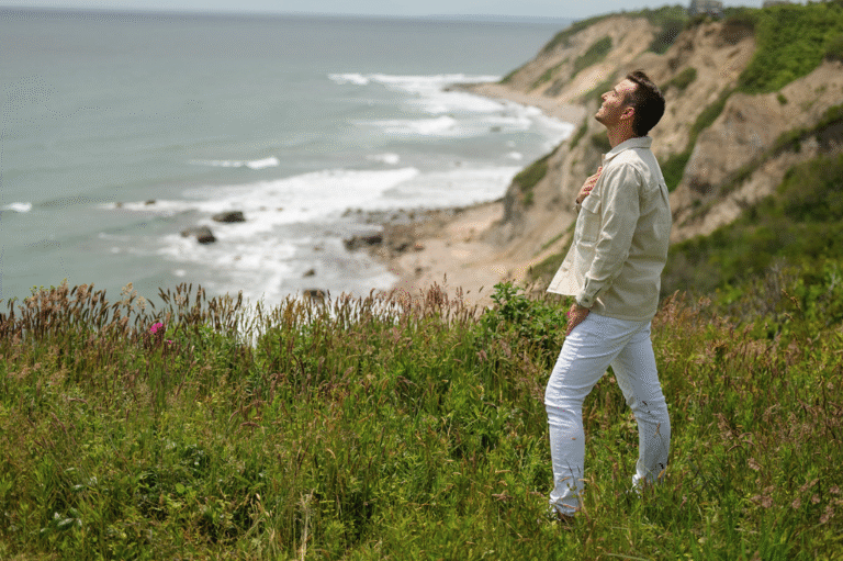 Matt Fraser, in a light shirt and white pants, stands on a grassy cliff by the ocean, looking upward with one hand on his chest.