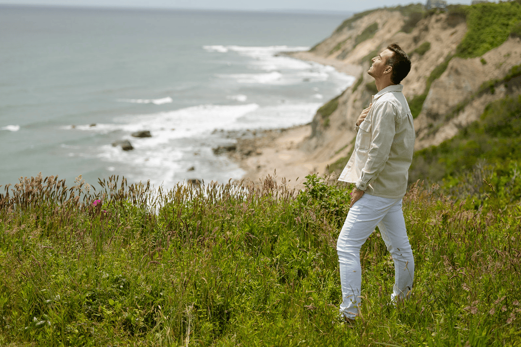 Matt Fraser, in a light shirt and white pants, stands on a grassy cliff by the ocean, looking upward with one hand on his chest.