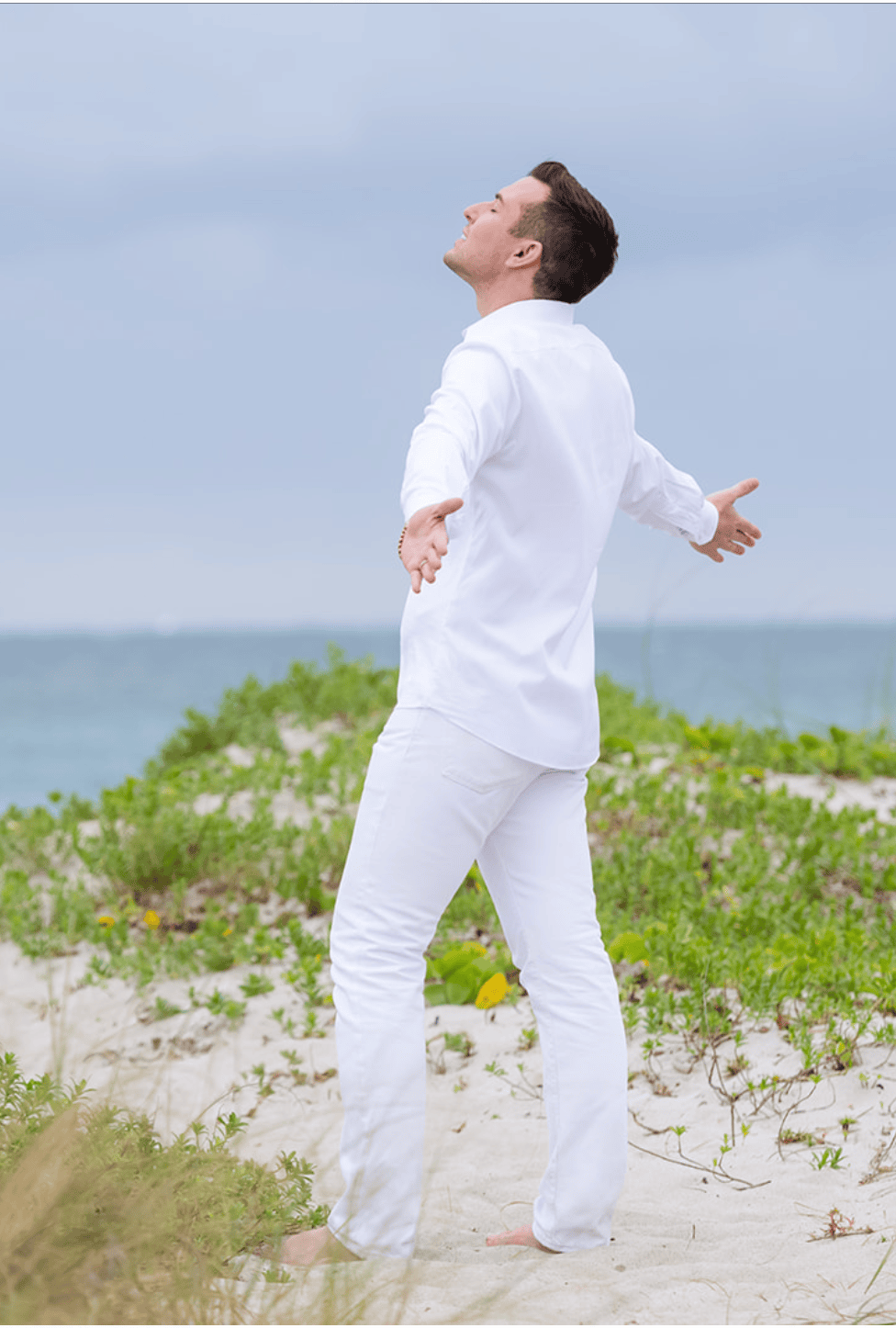 Matt Fraser in white clothes stands barefoot on a sandy beach with green plants, arms outstretched near the ocean under a cloudy sky.