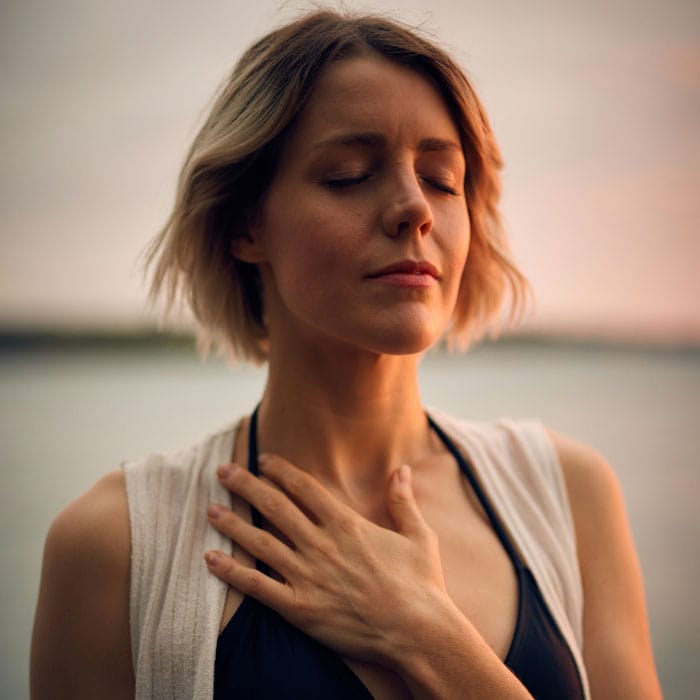 A woman with short hair stands outdoors, eyes closed, one hand on her chest, looking calm and serene.