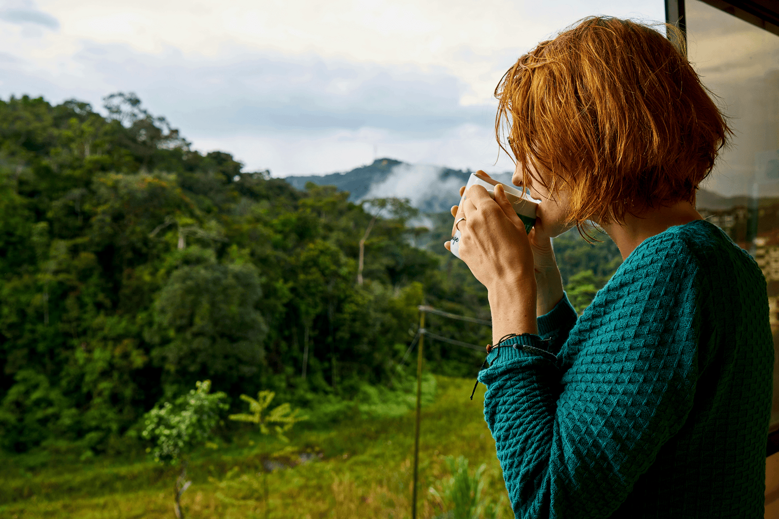 Matt Fraser with short red hair drinks from a cup while standing by a window overlooking a lush green forest and distant hills.