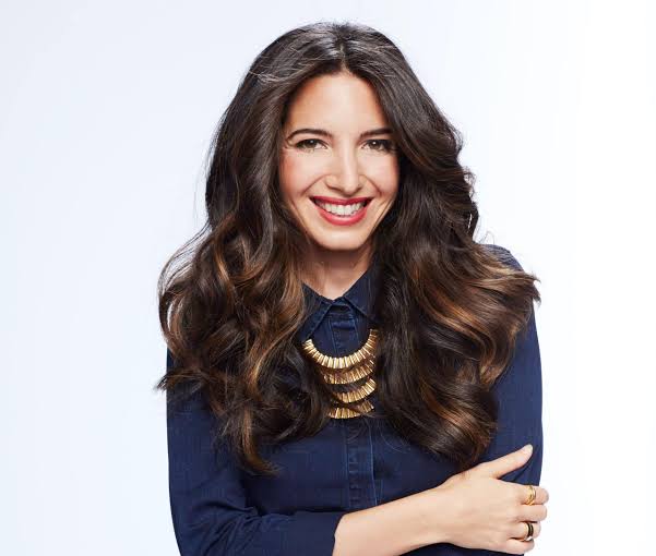 Certainly! Here’s the description with your requested instruction: Matt Fraser, with long wavy dark hair, wearing a dark blue shirt and gold statement necklace, smiling at the camera against a plain light background.