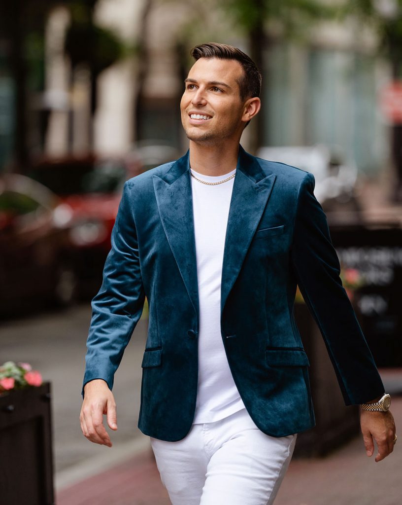 Matt Fraser, in a teal blazer and white shirt, walks outdoors on a city sidewalk, smiling and looking upward.