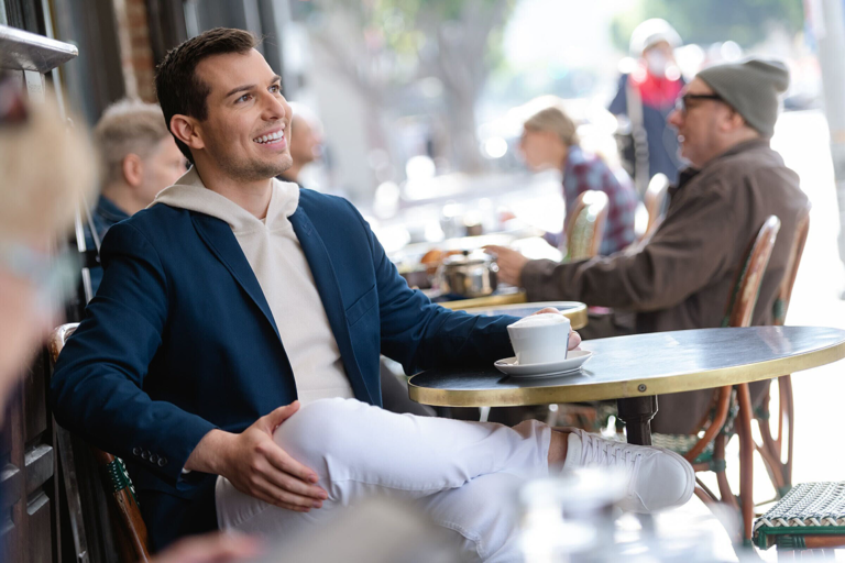 Matt Fraser, wearing a blue jacket and white pants, sits at an outdoor cafe table, holding a cup. Other people are seated in the background.