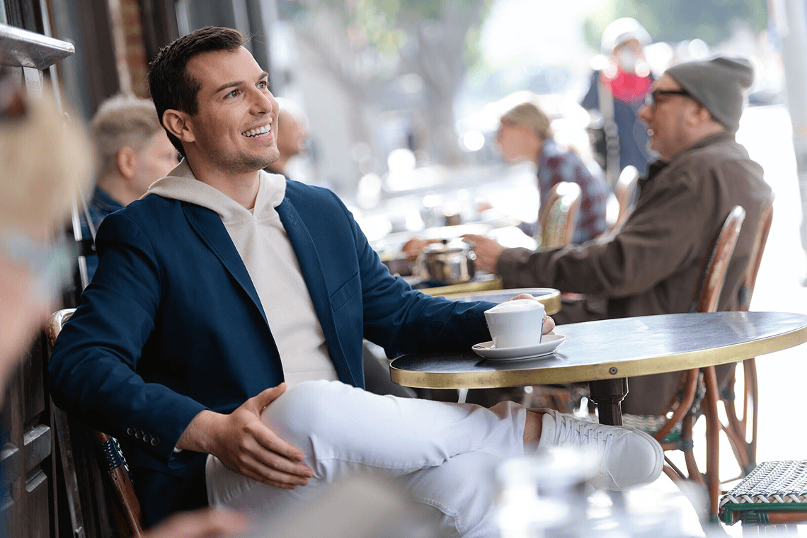 Matt Fraser, wearing a blue jacket and white pants, sits at an outdoor cafe table, holding a cup. Other people are seated in the background.