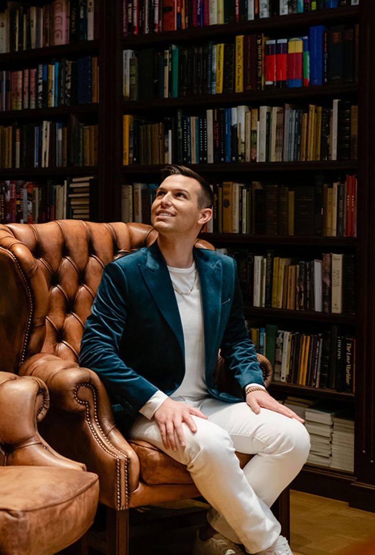 Matt Fraser, in a teal blazer and white pants, sits on a brown leather chair before a full bookshelf, looking up and smiling.