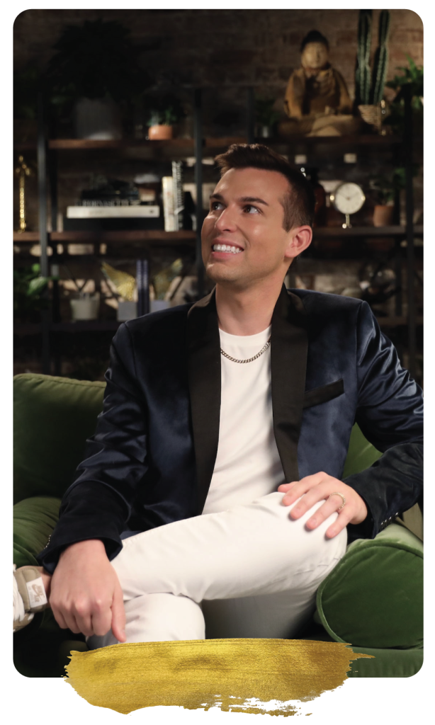 Matt Fraser in a dark blazer and white pants sits on a green chair, smiling upward, with shelves of plants and decor behind him.