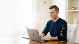 Matt Fraser sits at a wooden desk using a laptop, holding a pencil, with a microphone, notebook, and decor in the background.