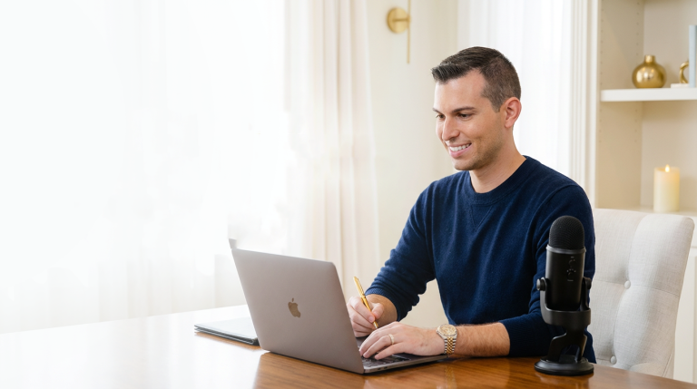 Matt Fraser sits at a wooden desk using a laptop, holding a pencil, with a microphone, notebook, and decor in the background.