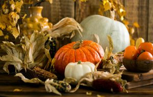 Assorted pumpkins, gourds, corn, and a wooden bowl of persimmons on a rustic table with dried leaves and a yellow vase behind.