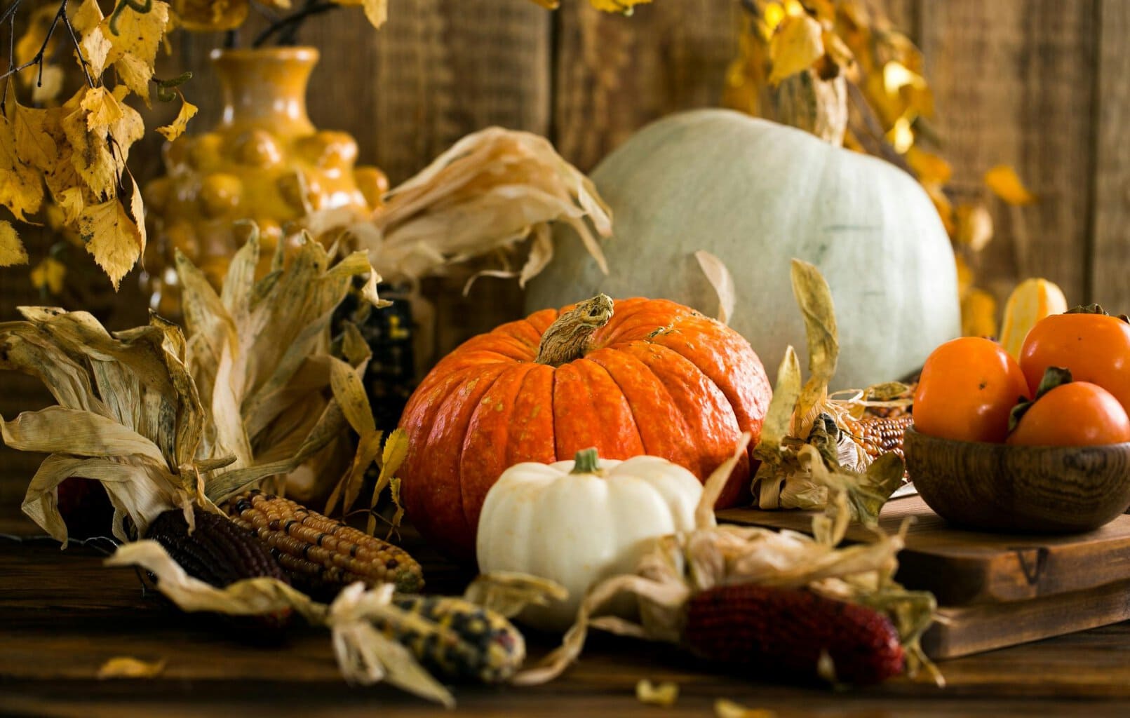 Assorted pumpkins, gourds, corn, and a wooden bowl of persimmons on a rustic table with dried leaves and a yellow vase behind.