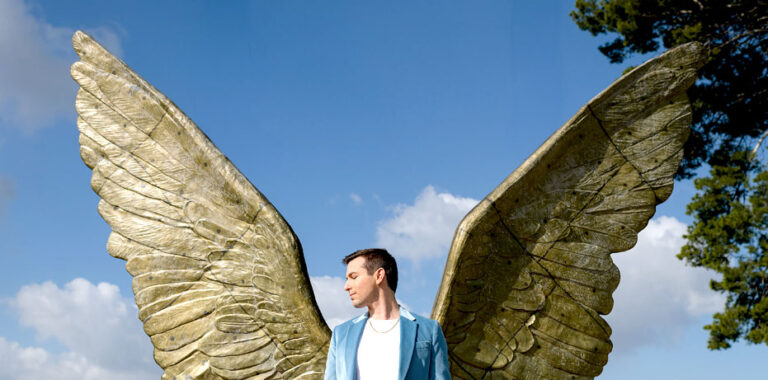Matt Fraser stands in front of a large, bronze-colored angel wings sculpture, creating the illusion that he has wings, with blue sky and clouds in the background.