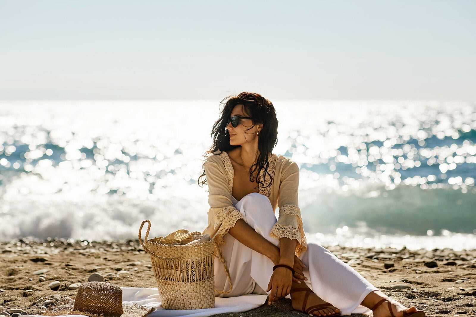A woman in sunglasses and light clothing sits on a sandy beach near a woven basket, with ocean and sunlight in the background.